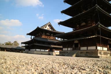 Wooden temple structures with a multi-tiered pagoda, standing on a ground of small stones under a blue sky.